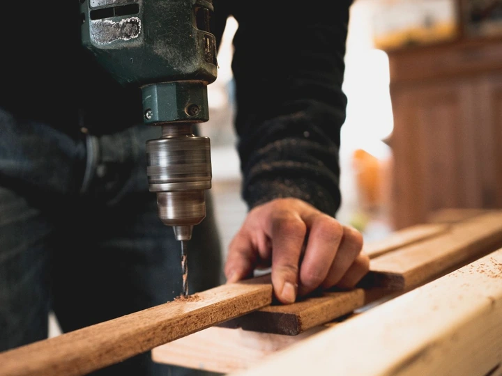 Une mèche de perceuse entre dans une latte de bois clair, tenue par une main, sur un établi couvert de sciure.